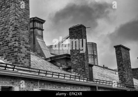 Schwarz-weiß Foto mit Schiefer Dach mit Ziegel Kamine im Vordergrund mit modernen Glasgebäude hinter und Wolken darüber hinaus. Stockfoto