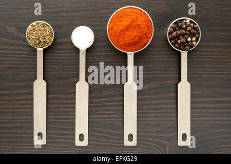 Pfefferkörner, Paprika, Thymian und Salz in Dosierlöffel auf einer dunklen Holzoberfläche Stockfoto
