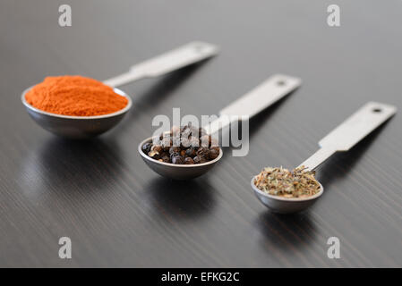 Pfefferkörner, Paprika und Thymian in Dosierlöffel auf einer dunklen Holzoberfläche Stockfoto