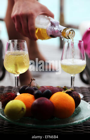 Die Hand eines Mannes gießt in zwei Gläser Champagner. Eine Platte mit exotischen Früchten ist in der Front. Stockfoto