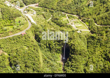 Aus der Vogelperspektive ist ein hoher, schmaler Wasserfall zu sehen, der auf einer steilen, bewaldeten Klippe in ein grünes Tal hinabstürzt. Die umliegende Landschaft ist dicht von lebhafter grüner Vegetation bedeckt Stockfoto