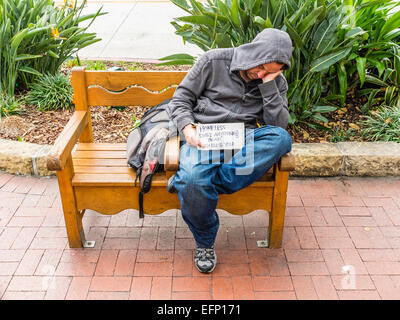 Ein Obdachloser schläft draußen auf einer Bank Bürgersteig auf der State Street in Santa Barbara, Kalifornien während einem bettelnden Namensschild. Stockfoto