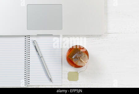 Winkel der Ansicht von oben geschossen des Desktops mit Notizblock, Stift, Laptop-Computer und Glas Tee mit Tee-Beutel im Inneren.  Querformat. Stockfoto