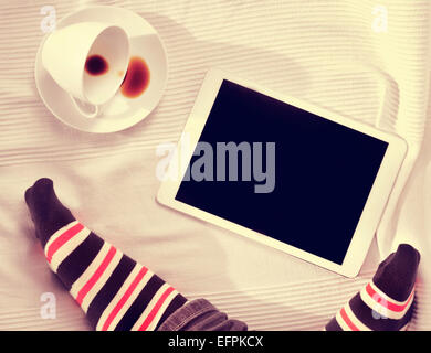 hoch-Winkel der Füße eines Mannes und einer Tasse Kaffee und einem Tablet-Computer auf einem Bett mit einer Bettdecke bedeckt Stockfoto