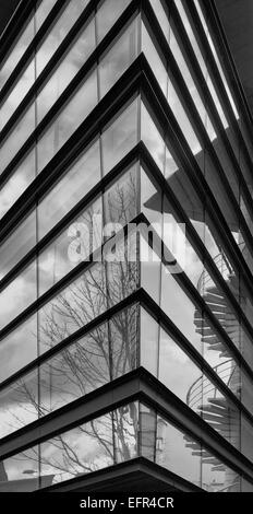 Schwarz / weiß Foto zeigt scharfe Ecke der Glasfassade mit Reflexionen von Wolken, Bäume und Wendeltreppe Stockfoto