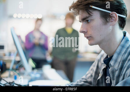 Ein junger Mann sitzt an einem Computer in einer Reparaturwerkstatt.  Zwei Personen im Hintergrund. Stockfoto