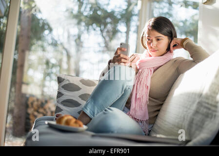 Frau sitzt auf einem Sofa, Blick auf ihr Handy, eine Platte mit einem Croissant vor ihr. Stockfoto