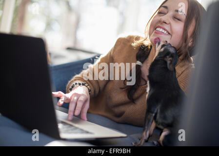 Frau auf einem Sofa Blick auf ihrem Laptop, lächelnd. Ein kleiner Hund, ihr Gesicht zu lecken. Stockfoto