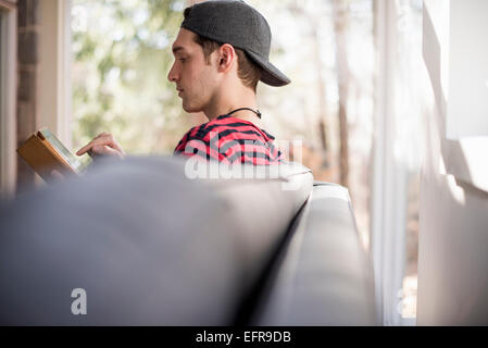 Mann trägt eine Baseball-Kappe nach hinten, auf einem Sofa sitzt, mit Blick auf eine digital-Tablette. Stockfoto