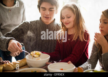 Vier Personen sitzen und stehen an einem Tisch, ein Mann mit Essen in eine Schüssel geben. Stockfoto