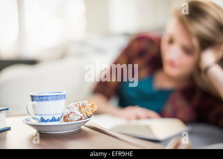 Junge Frau sitzt auf einem Sofa, ein Buch zu lesen. Eine Tasse und Untertasse mit einem Croissant auf einem Tisch. Stockfoto