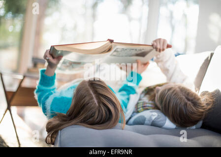 Zwei Mädchen auf einem Sofa auf dem Rücken liegend ein Buch lesen. Stockfoto