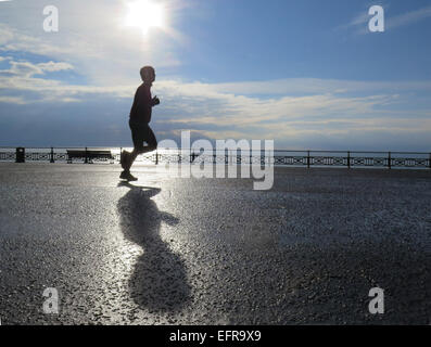 Ein männliche Jogger läuft Promenade Hove Rasen wirft seine Schatten in der frühen Morgensonne am glitzernden Meer. Stockfoto