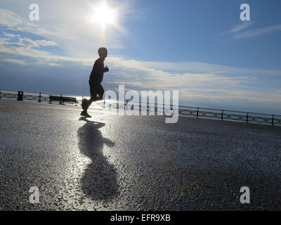 Ein männliche Jogger läuft Promenade Hove Rasen wirft seine Schatten in der frühen Morgensonne am glitzernden Meer. Stockfoto