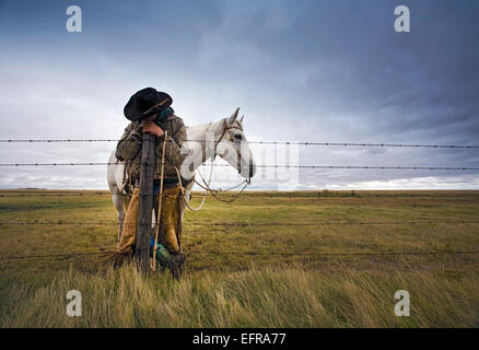 Ein Cowboy stehend schiefen post auf einen Zaun auf der Range. Ein graues Pferd hinter ihm. Stockfoto