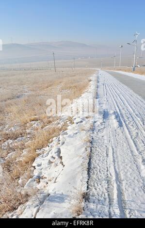 verschneite Straße mit Windkraftanlagen und Straßenlaternen powered by Sonnenkollektoren und Windkraftanlagen in Manzhouli Innere Mongolei, China. Stockfoto