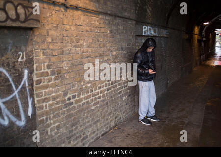 Young Asian man Unterstände Texte unter einer Eisenbahnbrücke, sich unter dem Deckmantel von Regen auf einem nassen Regentag in Whitechapel, East End von London, UK zu senden. Stockfoto