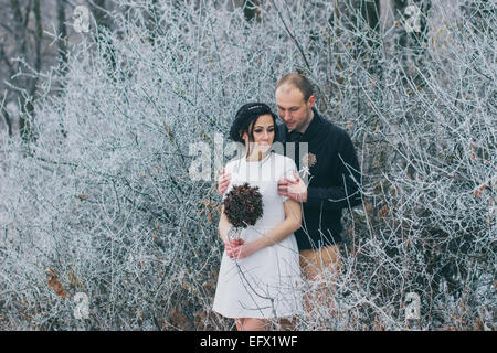 Bräutigam umarmt die Braut im tief verschneiten Winterwald; Braut Hochzeit Bouquet hält Stockfoto