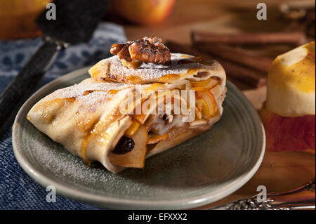 Apfelstrudel mit Walnüssen und Rosinen, serviert auf einem blauen Teller mit Äpfeln und Zimt im Hintergrund. Stockfoto