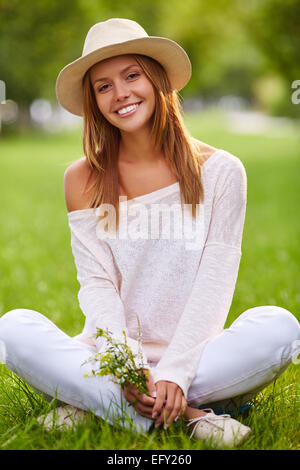 Stilvolle Mädchen in Hut und Casualwear auf Rasen im Park sitzen Stockfoto