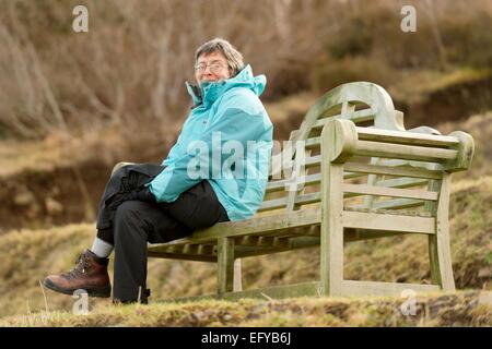 Frau in blauer Jacke sitzt in einer erhöhten große hölzerne Parkbank während eine Sportveranstaltung - Skye V Lovat, Shinty Spiel beobachten. Stockfoto