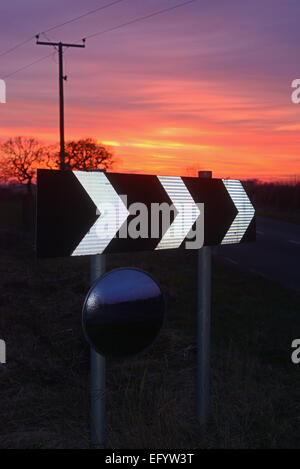 Sonnenuntergang von Warnzeichen der scharfen Kurve und kreisförmige Sicherheitsspiegel Fahrer Sicht auf Straße uk geben Stockfoto