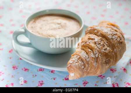 Schaumigen Kaffee in einem blauen Tasse und Untertasse auf einer floral Tischdecke mit Croissant Gebäck. Stockfoto