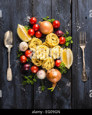 Pasta Tagliatelle mit Tomaten, Gemüse und Gewürze für Tomatensauce, Löffel und Gabel auf hölzernen dunkelblauem, Ansicht von oben Stockfoto