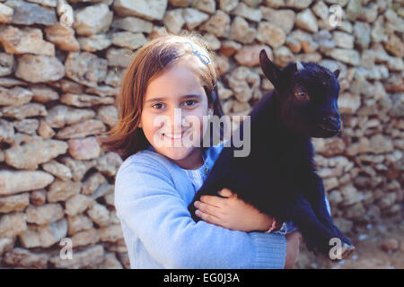 Lächelndes Mädchen, das an einer Wand steht und ein Ziegenbaby trägt Stockfoto