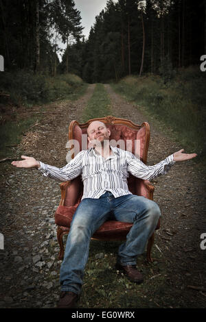 Schmierigen Mann in einem klassischen Vintage Stuhl in der Mitte einer Straße in den Wald. Zigarre und Cognac von einer Frau serviert bekommen. Stockfoto