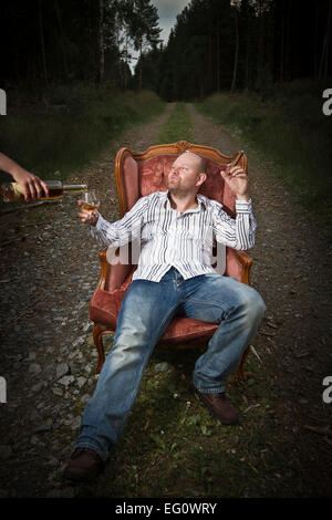 Schmierigen Mann in einem klassischen Vintage Stuhl in der Mitte einer Straße in den Wald. Zigarre und Cognac von einer Frau serviert bekommen. Stockfoto