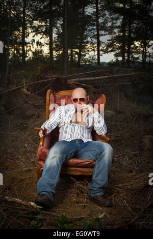 Schmierigen Mann in einem klassischen Vintage Stuhl in der Mitte eine Logging-Bereich. Zigarre rauchen und trinken Cognac in Wäldern. Stockfoto