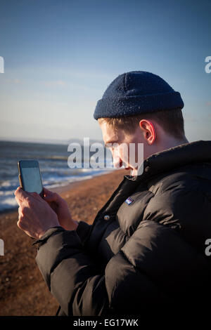 Junger Mann tragen 'Puffa' Jacke und wollene Mütze im Winter, fotografiert mit einem Ipad auf einem Kiesstrand. Stockfoto