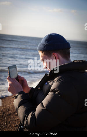 Junger Mann tragen 'Puffa' Jacke und wollene Mütze im Winter, fotografiert mit einem Ipad auf einem Kiesstrand. Stockfoto