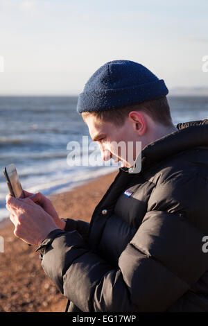 Junger Mann tragen 'Puffa' Jacke und wollene Mütze im Winter, fotografiert mit einem Ipad auf einem Kiesstrand. Stockfoto