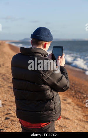 Junger Mann tragen 'Puffa' Jacke und wollene Mütze im Winter mit dem Fotografieren mit einem Ipad auf einem Kiesstrand. Stockfoto