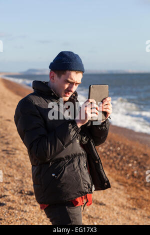 Junger Mann tragen 'Puffa' Jacke und wollene Mütze im Winter mit dem Fotografieren mit einem Ipad auf einem Kiesstrand. Stockfoto