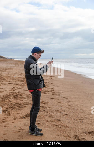 Junger Mann tragen 'Puffa' Jacke und wollene Mütze auf Chesil Beach im Winter mit dem Fotografieren mit einem Ipad. Stockfoto