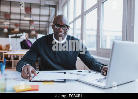 Porträt von zuversichtlich junger Mann an seinem Schreibtisch mit Laptop Papierkram zu tun. Afrikanische glücklich Blick in die Kamera bei der Arbeit. Stockfoto