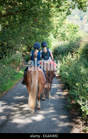 Zwei junge Mädchen reiten Ponys auf einer englischen Landstraße in Spätsommer-Sonnenschein. Stockfoto