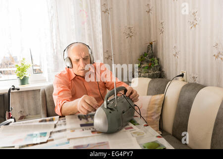 Alten Glatzkopf mit Kopfhörer hören von Kassetten-Player beim Sitzen am Tisch im Wohnzimmer mit Zeitungen. Stockfoto