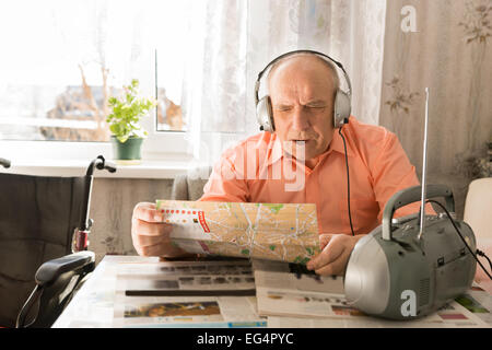 Aktualisiert alten Glatzkopf, sitzt am Tisch im Wohnzimmer, hören Musik von Radio mit Kopfhörer Gerät während Lesung hautnah Stockfoto