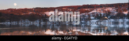 Schnee bedeckte Monk Coniston als die Sonne untergeht und der Mond rot färbt herauskommt, reflektierte im Seewasser Stockfoto