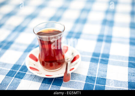 ISTANBUL, Türkei / Türkiye — Ein Glas türkischer Tee auf einer karierten blau-weißen Tischdecke in Istanbul, Türkei. Türkischer Tee wird traditionell in kleinen Gläsern serviert, die so geformt sind, auf einem kleinen Teller. Es ist ein schwarzer Tee, der ohne Milch, manchmal aber mit Rübenzucker eingenommen wird. Stockfoto