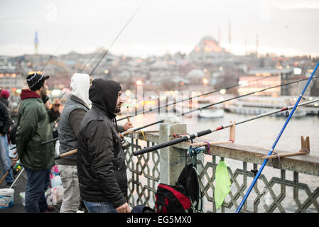 ISTANBUL, Türkei / Türkiye — Fischer säumen den Rand der Galata-Brücke mit ihren Linien über die Seite zum Goldenen Horn. Die Galata-Brücke, die das Goldene Horn überspannt und Eminonu mit Karakoy verbindet, ist eine zweistöckige Brücke, die Straßen-, Straßenbahn- und Fußgängerverkehr auf der obersten Ebene mit Restaurants und Bars auf der darunter liegenden Ebene abwickelt. In der Ferne befindet sich die Suleymaniye-Moschee. Stockfoto