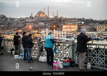 ISTANBUL, Türkei – Fischer säumen den Rand der Galata-Brücke mit ihren Leinen über der Seite in das Goldene Horn. Die Galata-Brücke erstreckt sich über das Goldene Horn und verbindet Eminonu mit Karakoy. Sie ist eine zweistöckige Brücke, die den Straßen-, Straßenbahn- und Fußgängerverkehr auf der obersten Ebene mit Restaurants und Bars auf der unteren Ebene regelt. In der Ferne ist die Suleymaniye-Moschee. Stockfoto