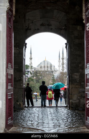 ISTANBUL, Türkei – Besucher mit Regenschirmen passieren das Kaisertor (Bâb-ı Hümâyûn) am Topkapi-Palast bei Regenfällen, wobei die Kuppeln und Minarette der Blauen Moschee durch den Torbogen sichtbar sind. Das 1478 erbaute Tor diente über vier Jahrhunderte lang als Haupteingang zum Palast. Die Szene zeigt zwei der bedeutendsten osmanischen Denkmäler Istanbuls in einem einzigen Bild. Stockfoto