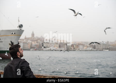 Galata Tower Seagulls Eminonu Waterfront Istanbul // ISTANBUL, Türkei - Ein Mann ernährt Möwen am Eminonu Waterfront entlang des Goldenen Horns, wobei der historische Galata Tower im Hintergrund sichtbar ist. Der Galata-Turm, 1348 von den Genuesen erbaut, ist ein mittelalterlicher Steinturm, der sich 67 Meter (220 Fuß) über dem Galata-Viertel in Beyoglu erhebt. Der Stadtteil Eminonu ist eines der geschäftigsten Geschäftsviertel Istanbuls und liegt auf der europäischen Seite der Stadt, wo das Goldene Horn auf den Bosporus trifft. Das Hafengebiet ist bekannt für seine Fährterminals, Fischmärkte und Straßenverkäufer. Möwen sind com Stockfoto