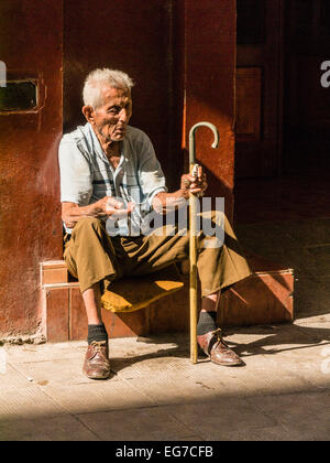 Eine ältere männliche kubanischen Bettler sitzt auf einer Tür Stoop hielt seine Hand für Geld mit einem Stock in Havanna, Kuba. Stockfoto