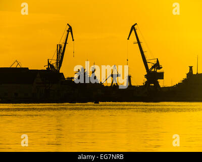 Containerschiff Krane Silhouette bei Sonnenaufgang gegen einen orangefarbenen Himmel und Wasser im Hafen von Havanna, Kuba. Stockfoto